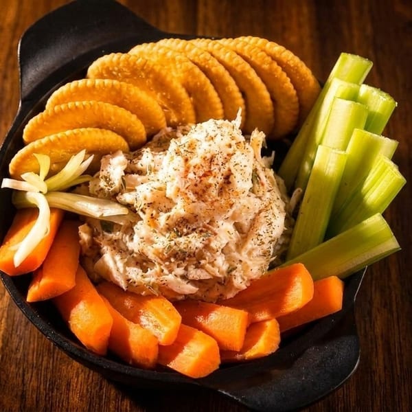 A black bowl of crab dip with crackers, celery sticks, and carrot sticks on a wooden table.