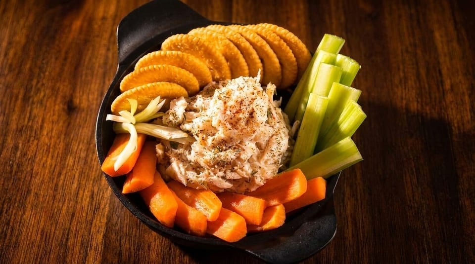 A black bowl of crab dip with crackers, celery sticks, and carrot sticks on a wooden table.